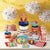 Image of a birthday party table with carnival and circus themed party supplies, there are popcorn buckets, cake decorations, napkins, paper plates and a table centrepiece.