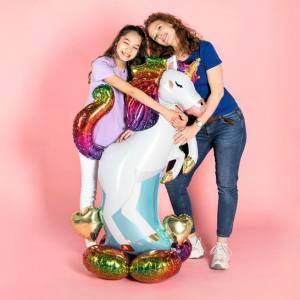 Image of a woman and a girl hugging a self standing unicorn balloon